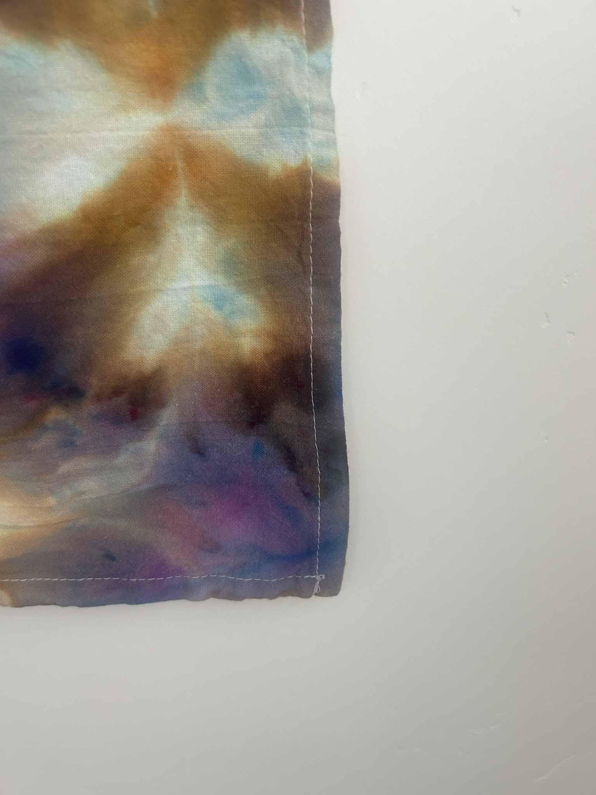 Close-up of ice dye tea towel corner and hem showing gold, purple, and blue color blending