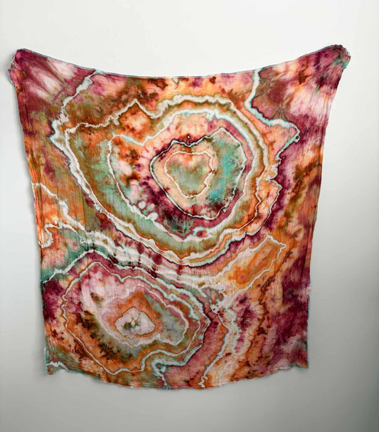 Square ice dyed bamboo gauze swaddle hanging on a white wall, showing two large geodes in raspberry and tangerine orange with sage green and aqua centers and white crackle lines. __etsy:8672499383