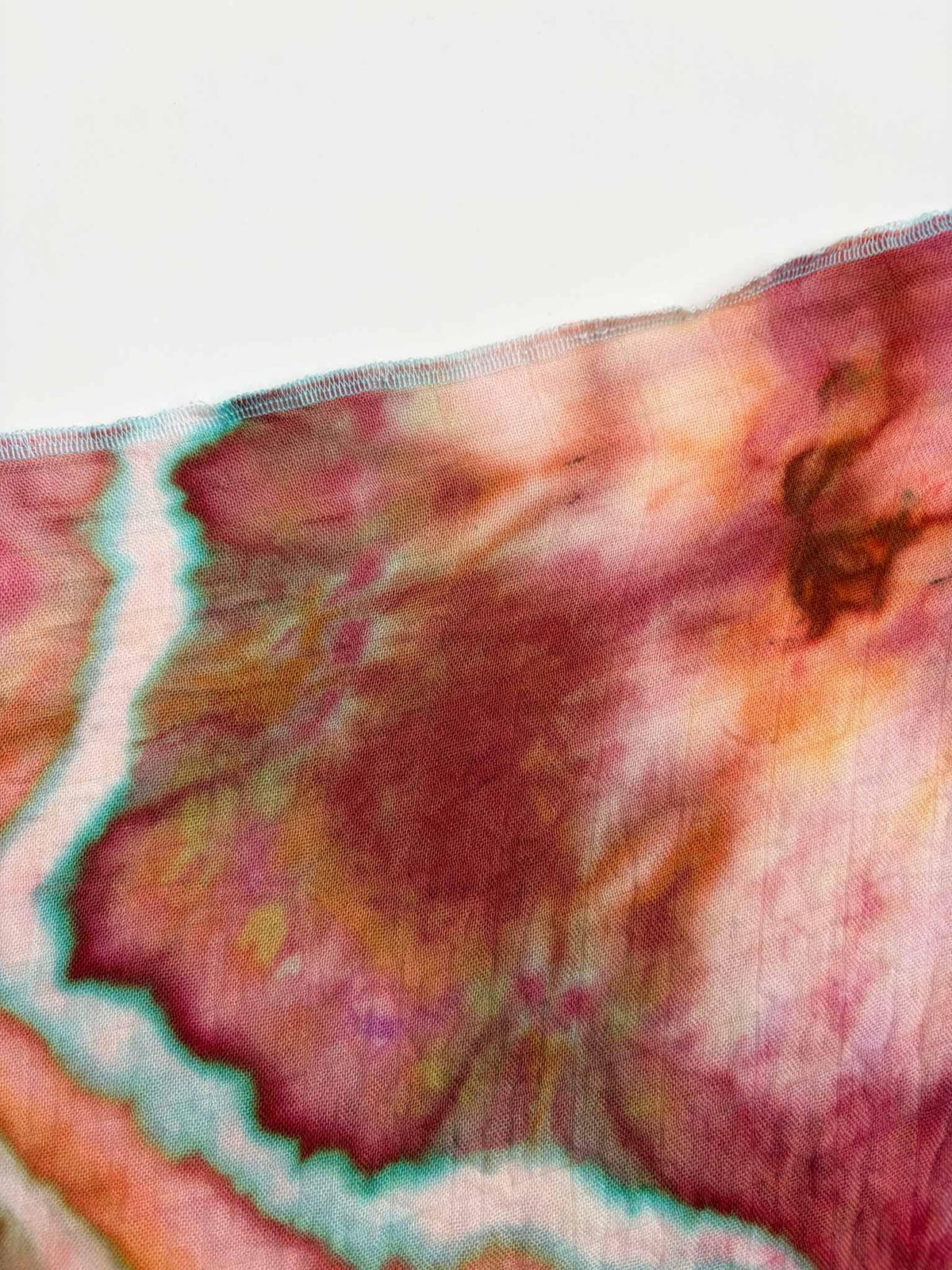Edge of a raspberry and coral ice dyed gauze swaddle showing the sheer weave, a white line edged in teal, and an aqua overlocked hem. __etsy:8672499385