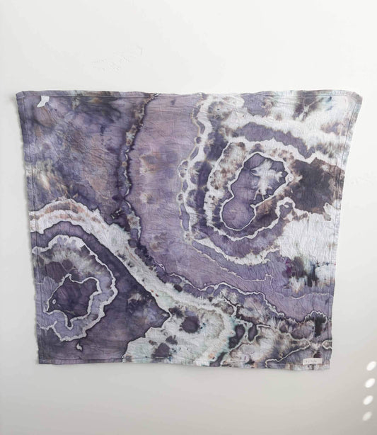 Square ice dyed tea towel hanging on a white wall, showing a geode pattern in lavender and slate purple with wide white bands, charcoal spots, and tan speckles. __etsy:8672439205
