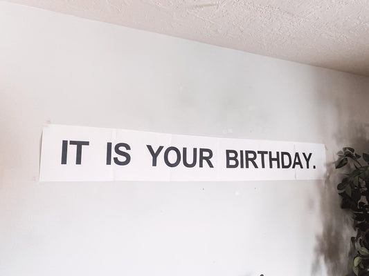 A white banner with black text that reads "IT IS YOUR BIRTHDAY." The banner is hanging on a white wall. Party item for The Office fans. Dwight Shrute inspired party decor.