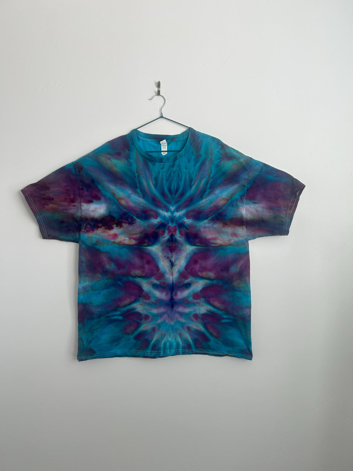 Front view of a mirrored ice dye Hanes 2XL tee in teal and plum hanging on a hanger against a white wall
