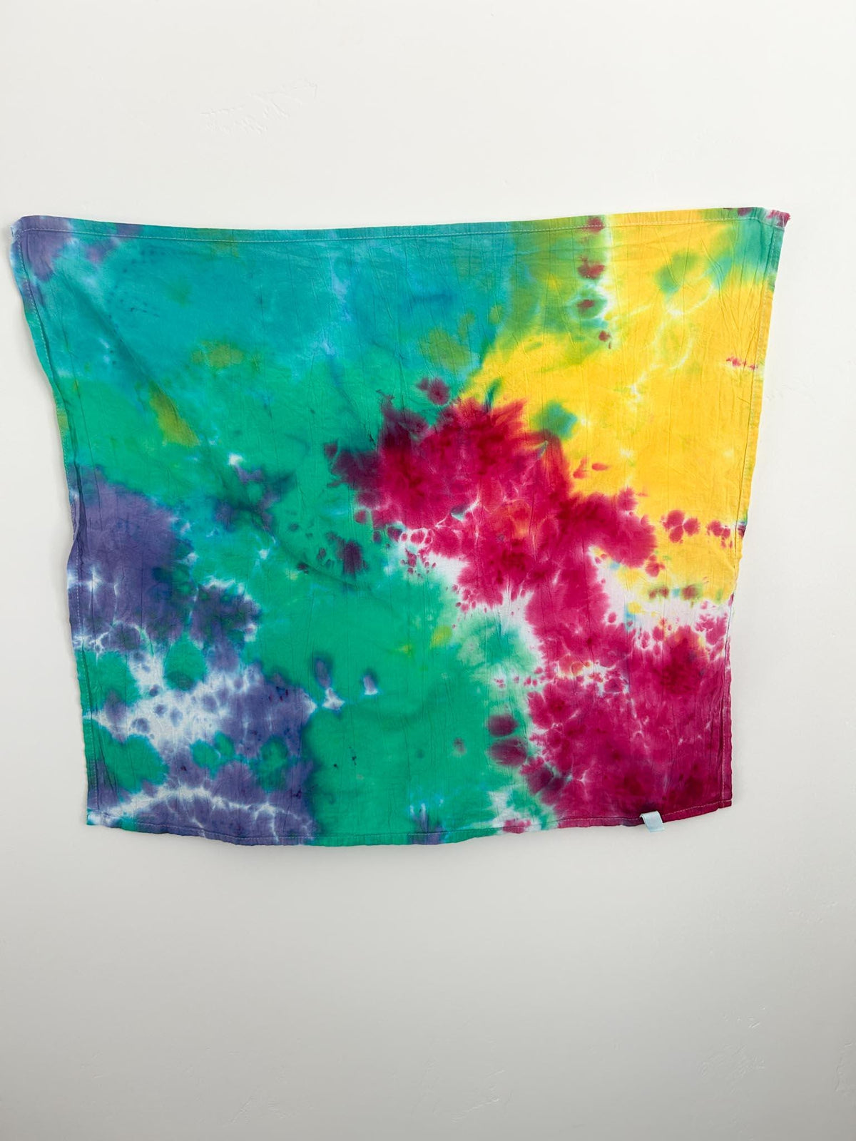 Full view of handmade rainbow tie dye flour sack tea towel hanging flat on white wall, 28 by 28 inches