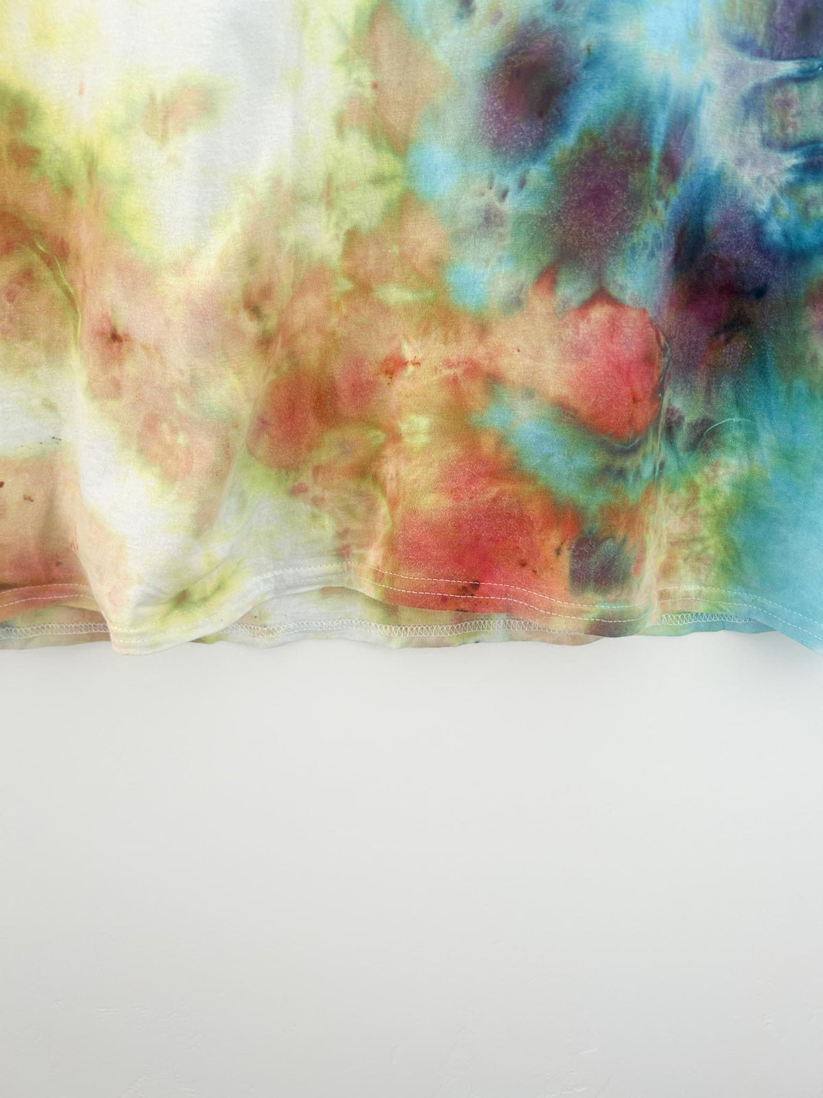 Bottom hem detail of ice dye tee showing coral, rust, yellow, and turquoise watercolor pattern