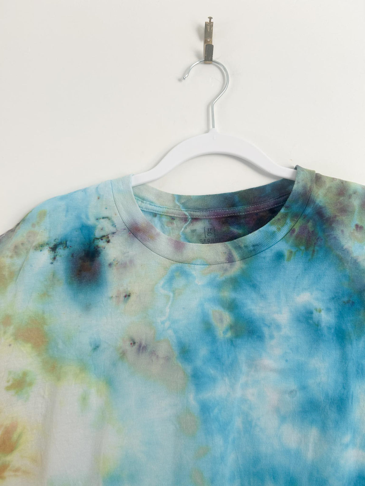 Crew neckline and shoulder of multicolor tie dye t shirt on white hanger showing teal and earth tone bleeds