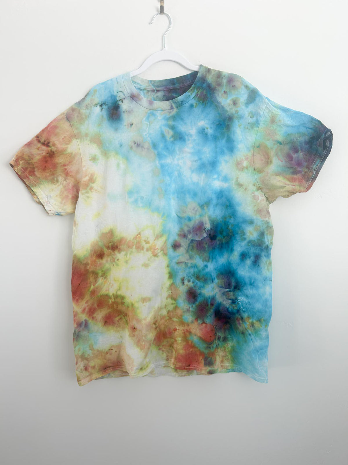 Front of handmade ice dye t shirt in size XL on white hanger, featuring a watercolor blend of sky blue, rust orange, chartreuse, and plum