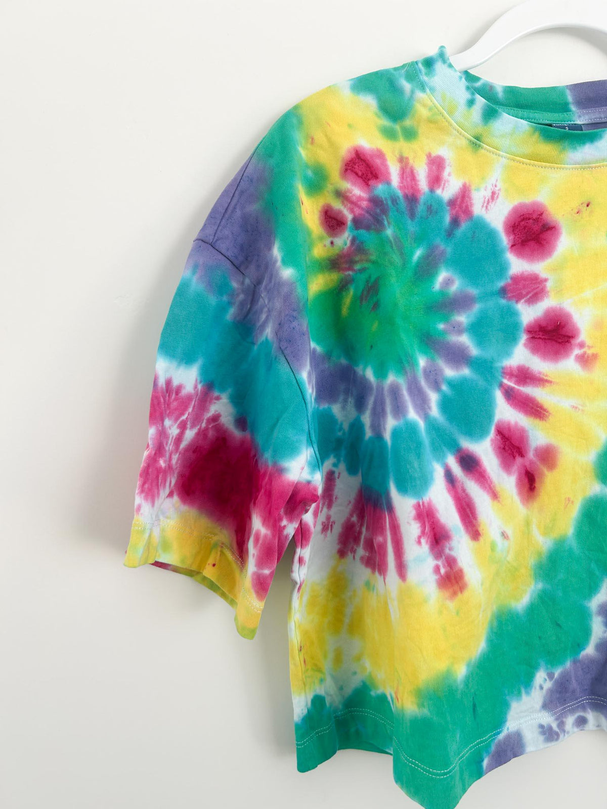 Side angle of handmade rainbow spiral tie dye crop top on white hanger with sleeve detail