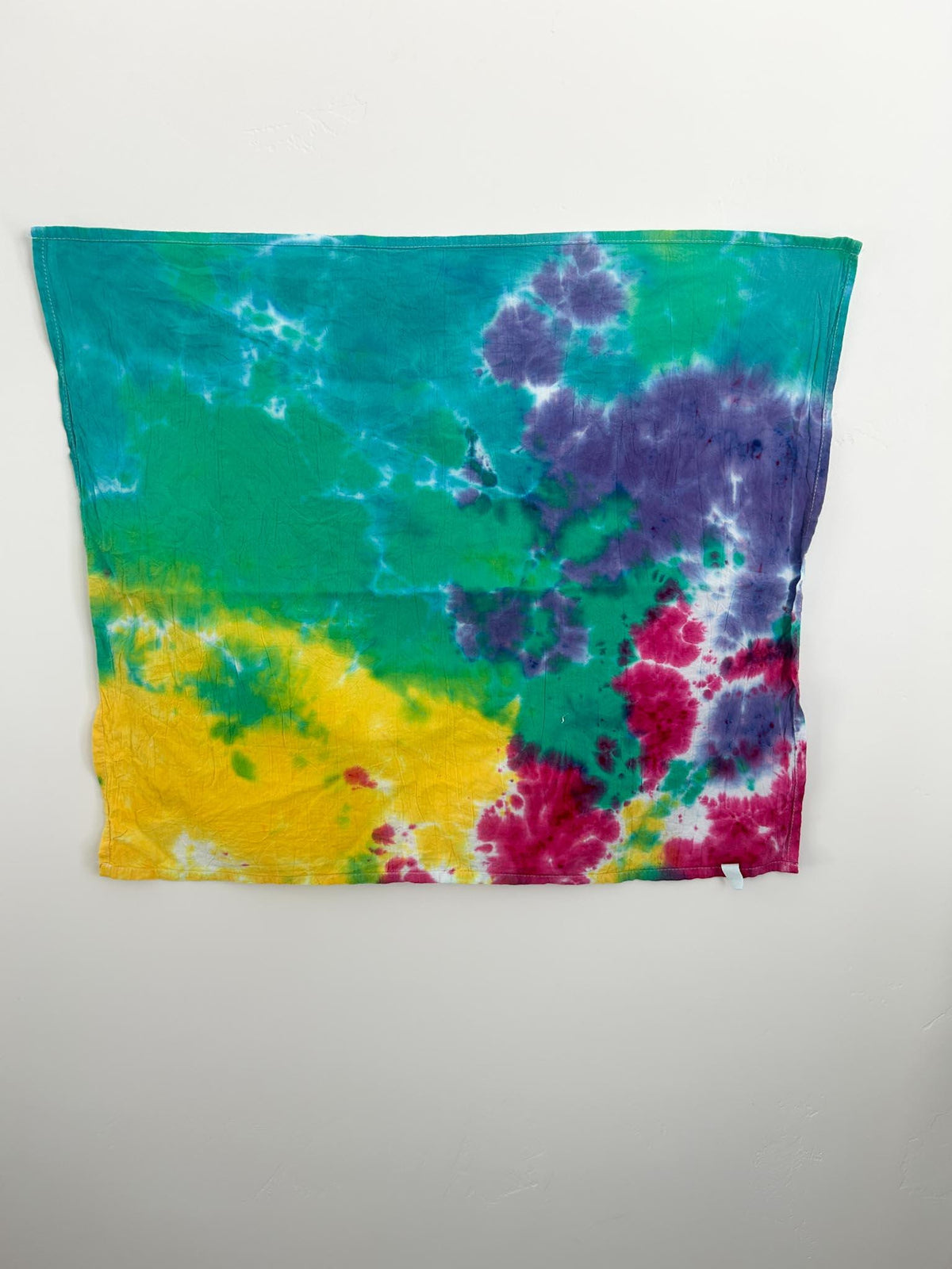 Full view of handmade rainbow tie dye flour sack tea towel hanging flat on white wall, 28 by 28 inches