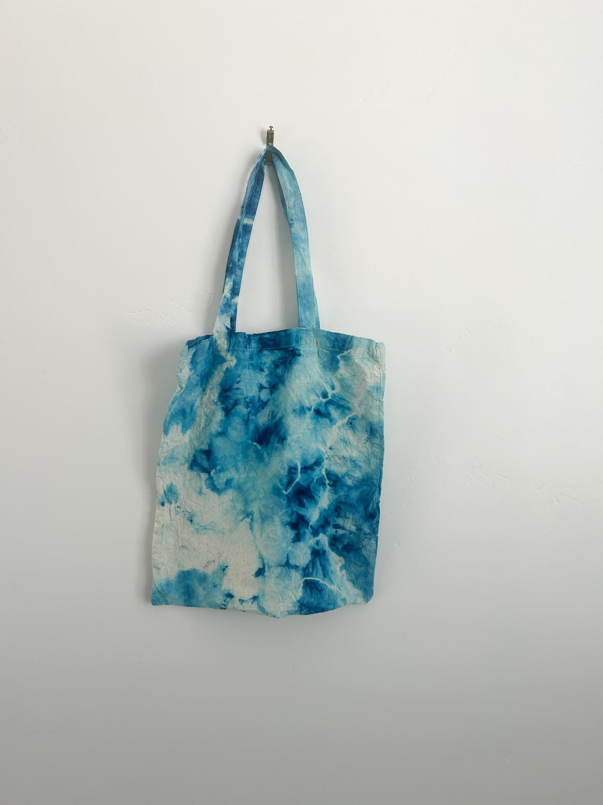 Front of handmade blue ice dye canvas tote bag hanging on white wall, sky blue and cobalt watercolor pattern