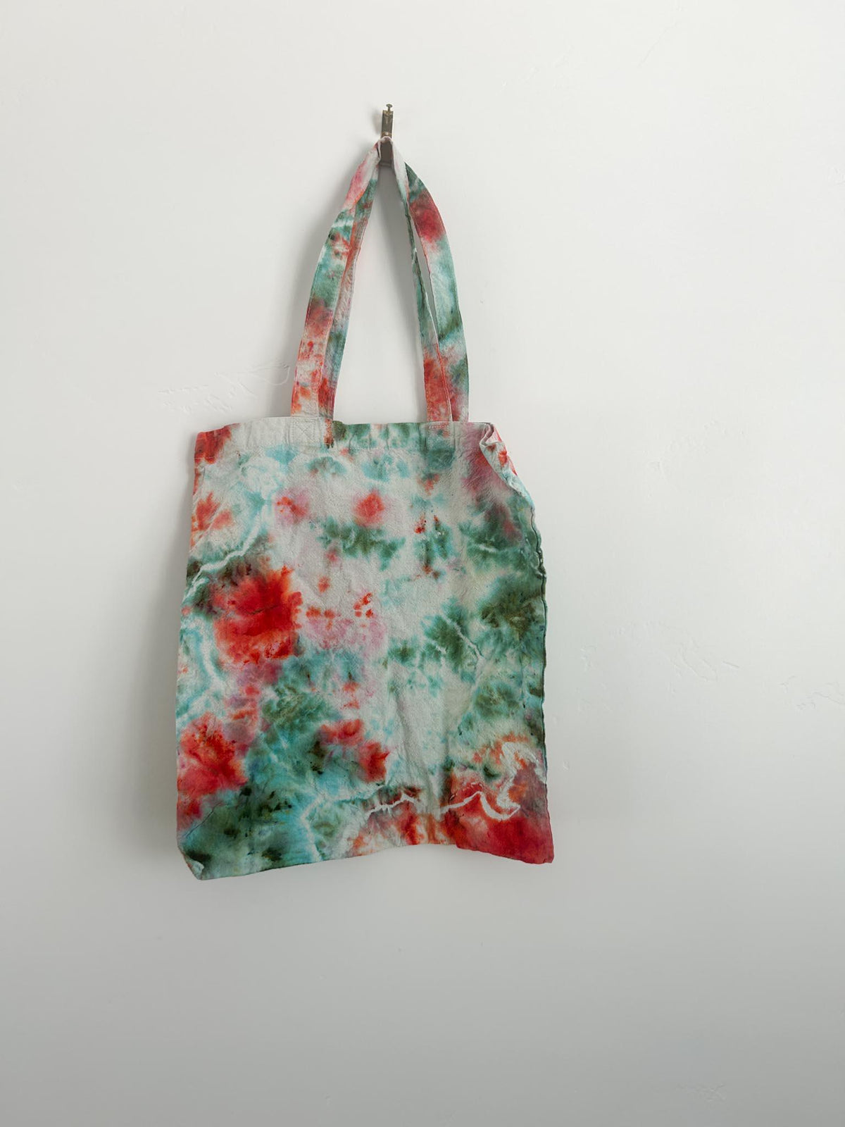 Handmade coral and teal ice dye canvas tote bag hanging on white wall, soft watercolor bloom pattern