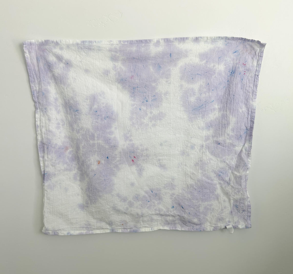 Wisteria ice dye tea towel in soft lavender on white cotton, with a cloudy purple pattern and small blue and pink speckles, hanging flat against a wall.