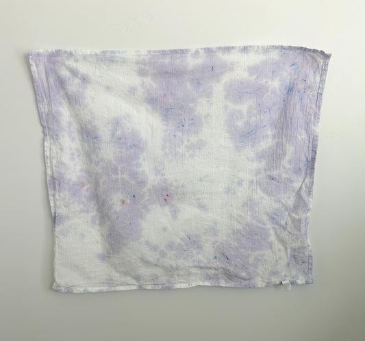 Wisteria ice dye tea towel in soft lavender on white cotton, with a cloudy purple pattern and small blue and pink speckles, hanging flat against a wall.