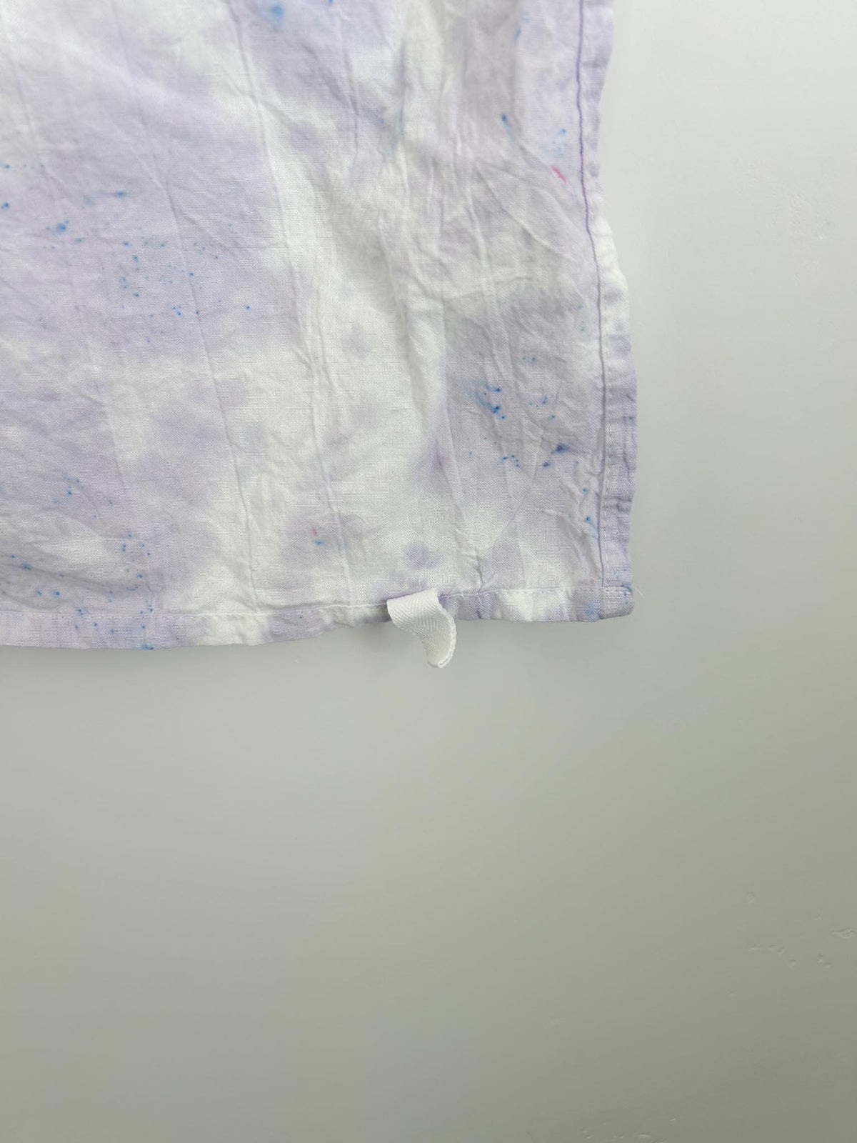 Close-up of the corner of a lavender ice dye cotton tea towel, showing the white hang loop and the blue and pink dye speckles in the fabric.