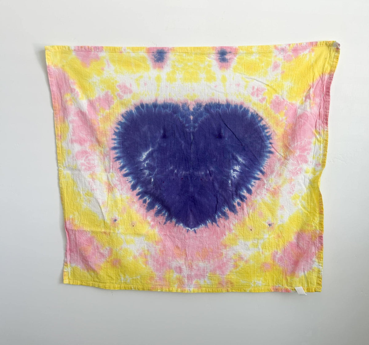 Hand dyed cotton tea towel with a purple heart pattern on a pink and yellow background, hanging flat.