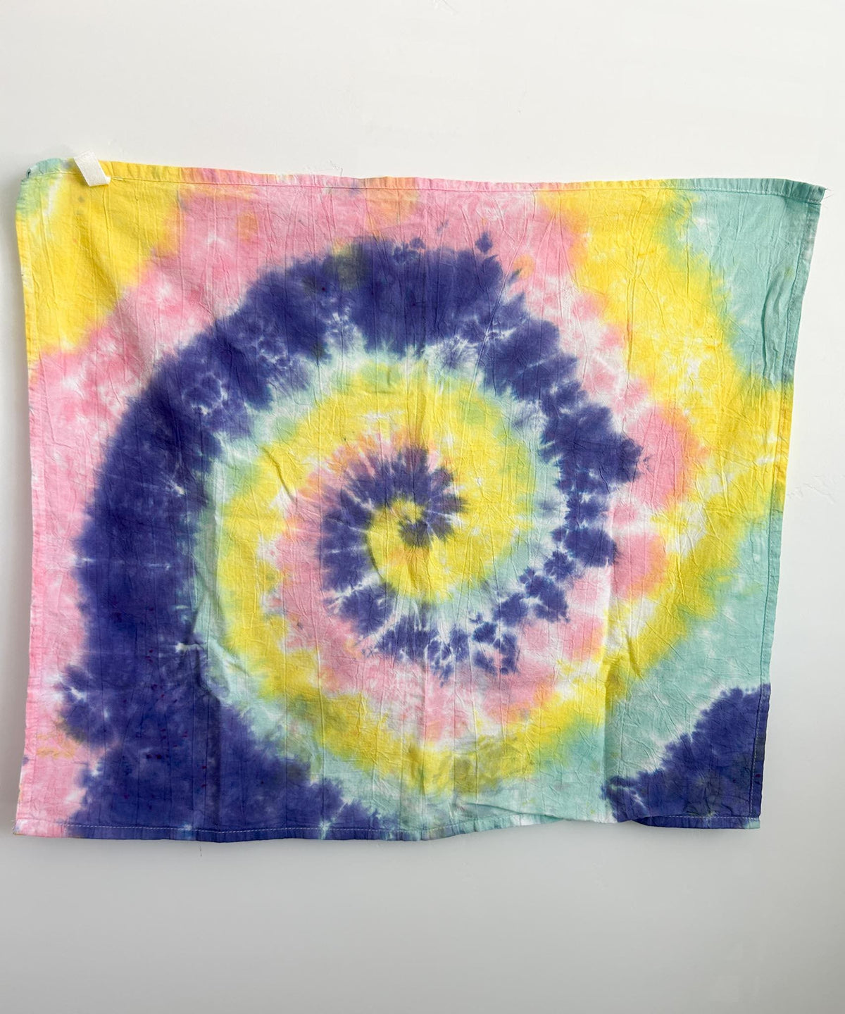 Pastel spiral tie dye cotton tea towel in purple, yellow, pink, and mint, hanging flat against a white wall.