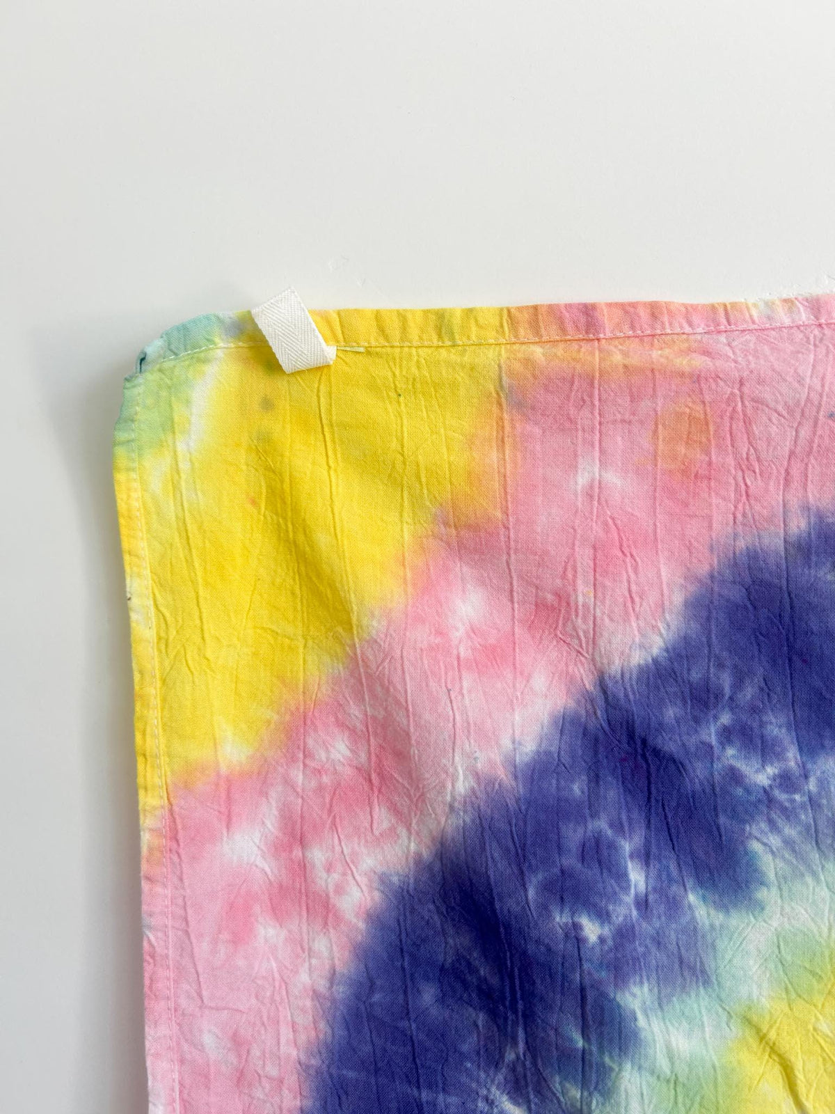Close-up corner of a pastel spiral tie dye tea towel showing the white hang loop and color gradient.