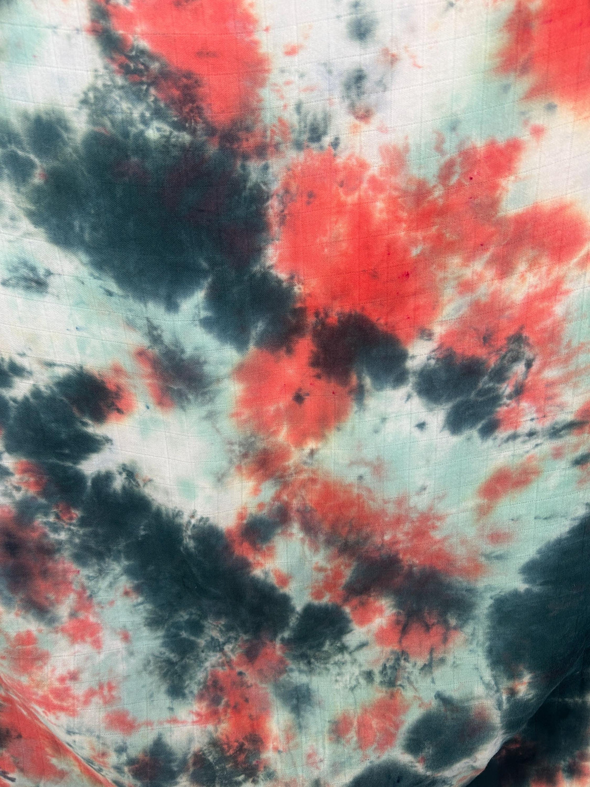 Close-up of a tie dye swaddle blanket showing bold coral red and charcoal pools on a mint background.