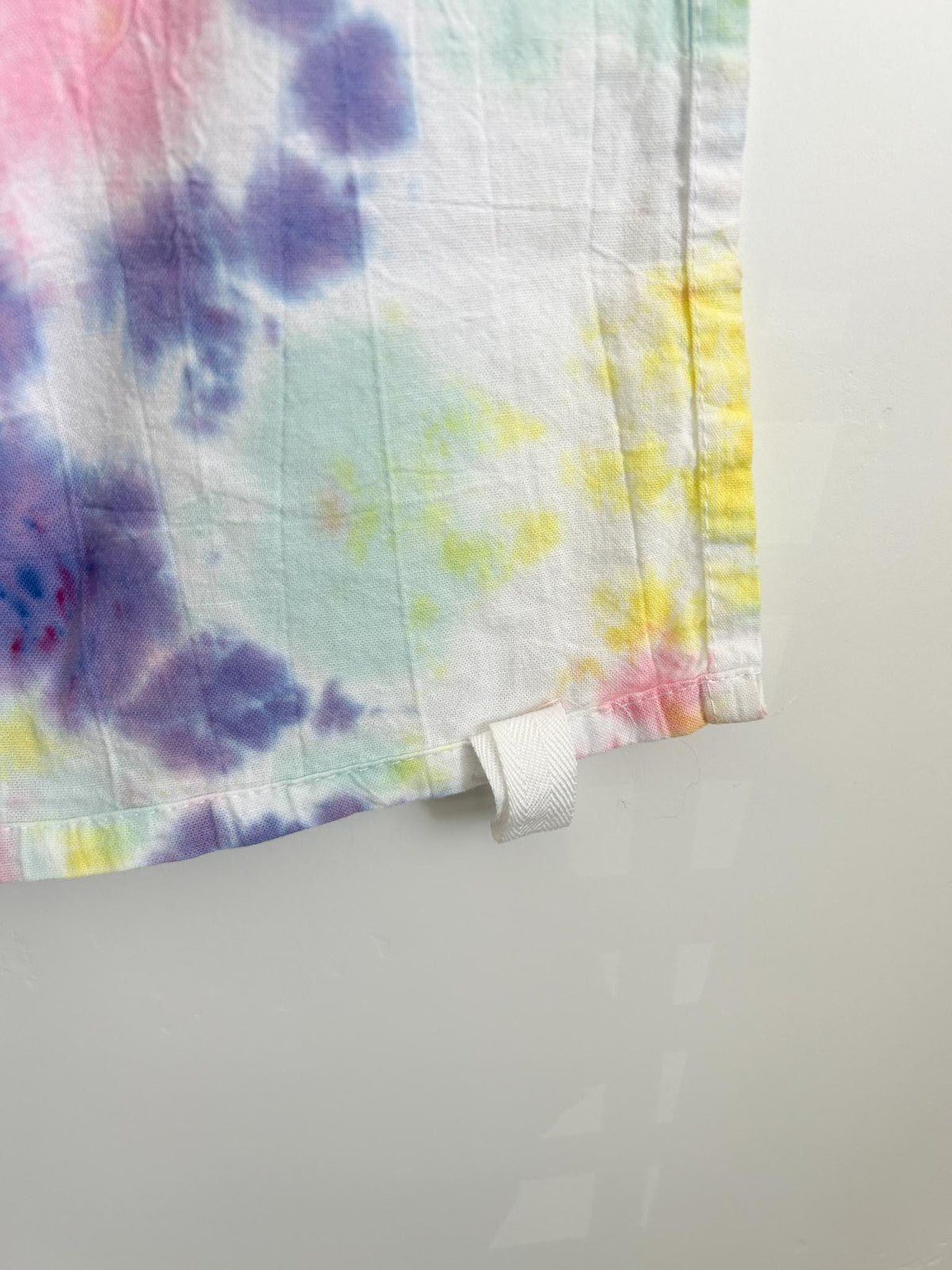 Close-up corner of a pastel scrunch tie dye tea towel showing the white hang loop and soft colors.