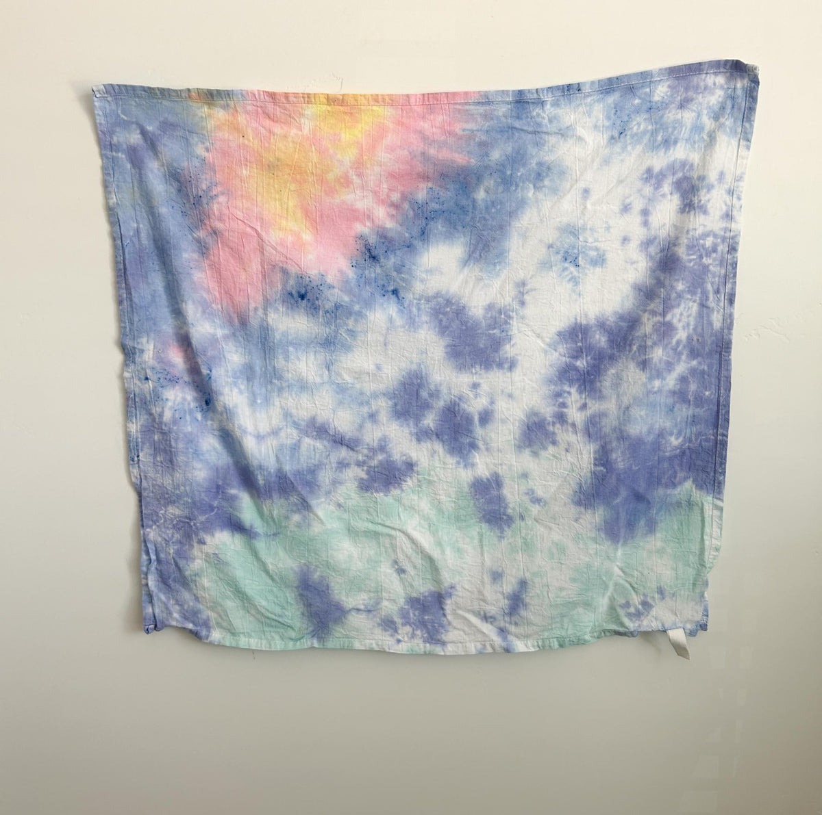 Soft pastel tie dye cotton tea towel in periwinkle, pink, peach, and mint, hanging flat against a white wall.