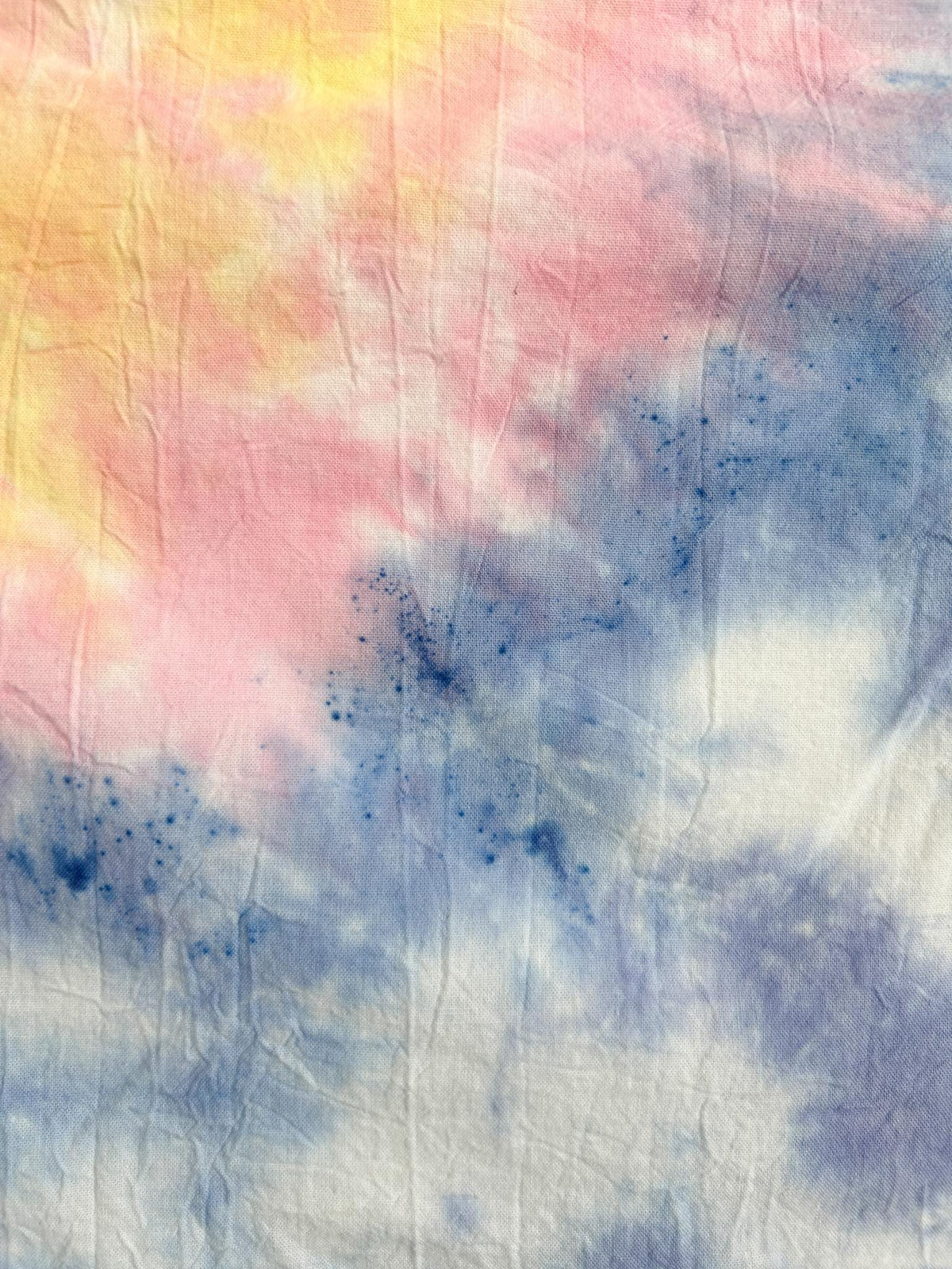Close-up of a pastel tie dye tea towel showing pink, yellow, and periwinkle blending in a watercolor pattern.