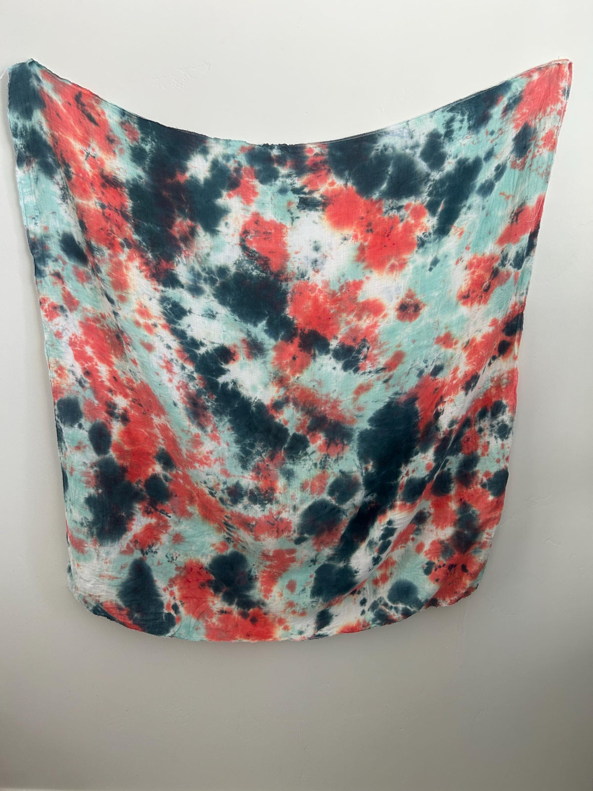 Hand dyed bamboo gauze swaddle blanket in coral red, charcoal, and mint, hanging flat against a white wall.