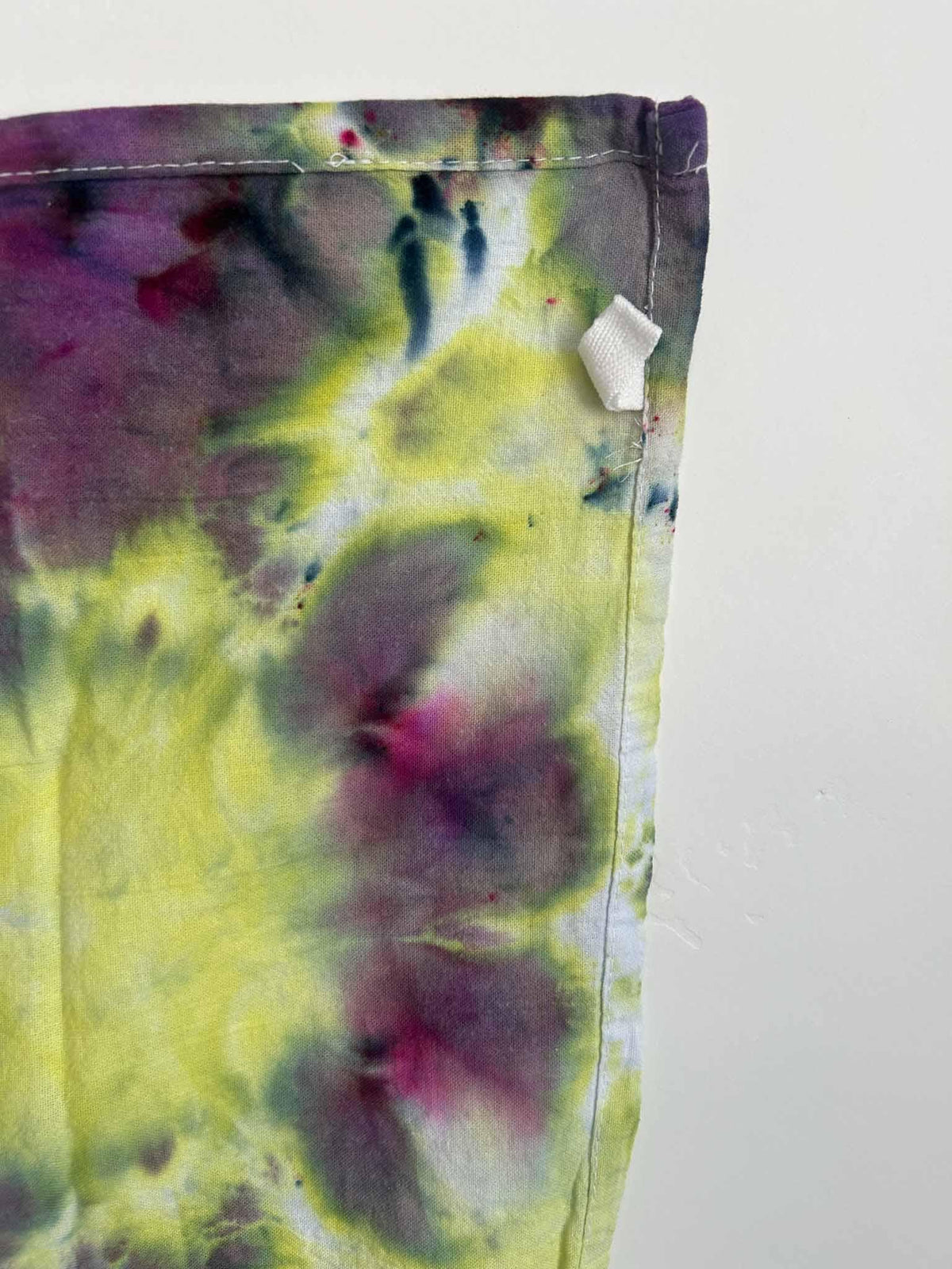Detail of a hand dyed yellow and purple tea towel showing the hemmed edge and white corner hang loop.