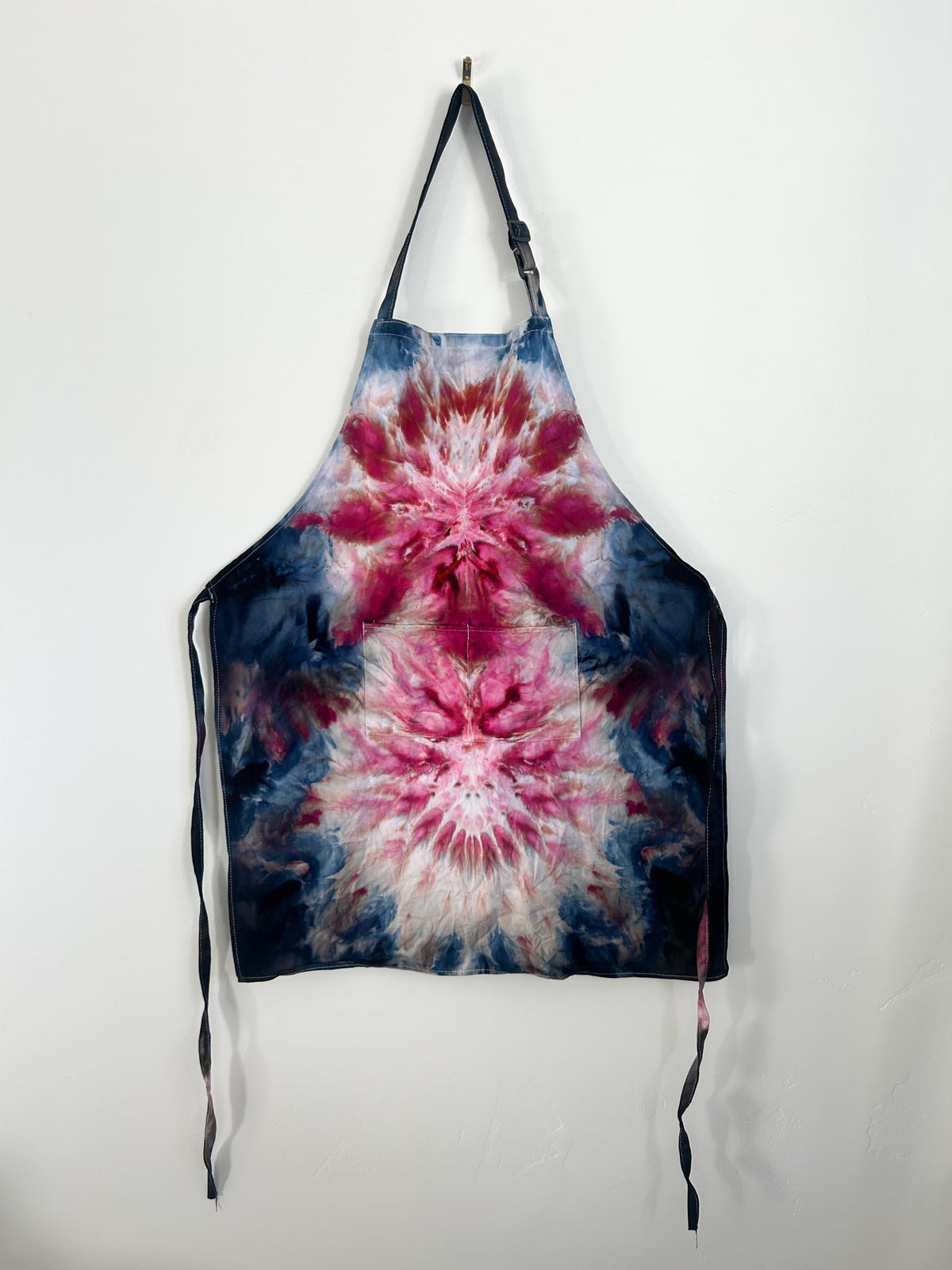 Navy burgundy tie dye apron hanging on hook, full view with adjustable strap and pocket