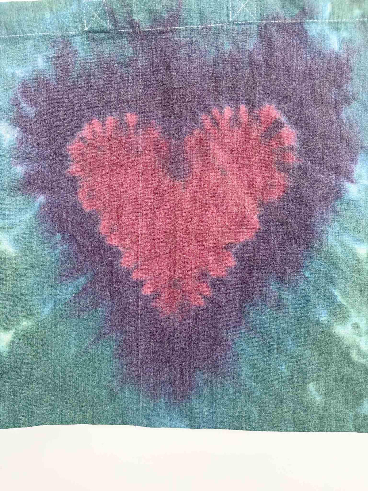 Close up of crimson heart tie dye pattern on cotton canvas tote bag in green and purple