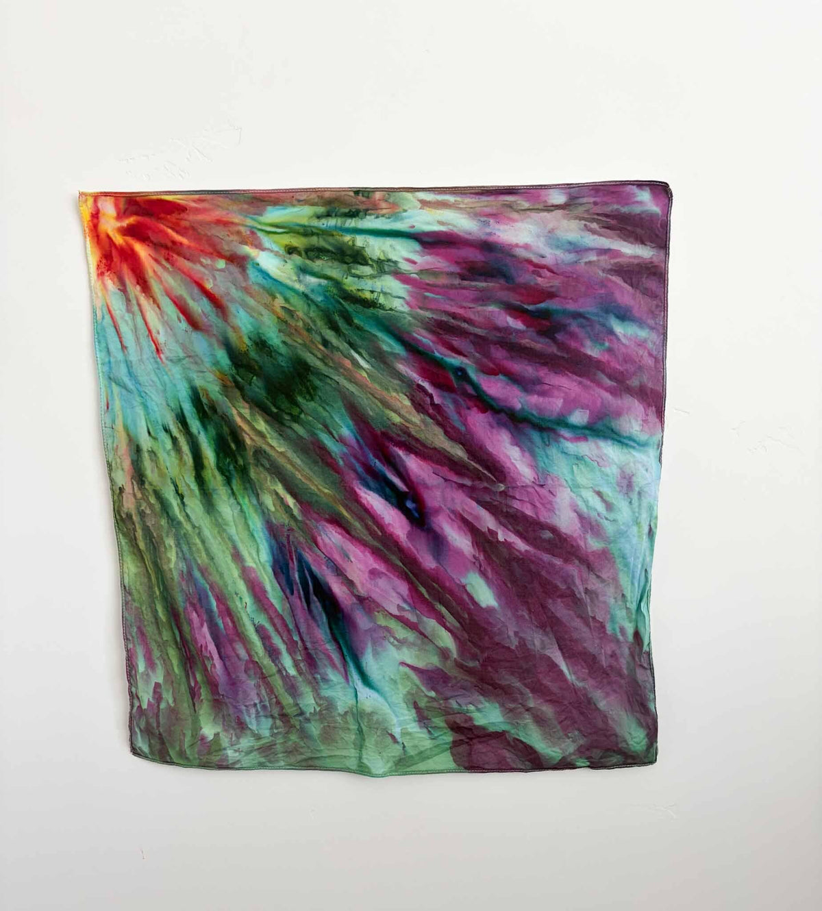 22x22 inch ice dyed cotton bandana with orange sunburst corner radiating into green and magenta fan fold pattern hanging on a wall