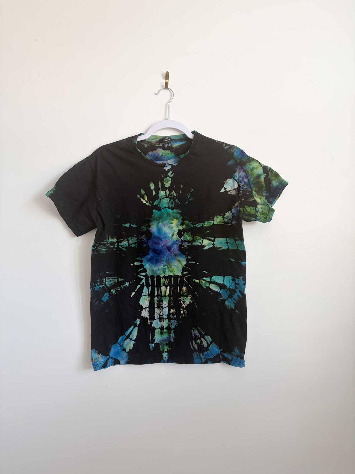 Small black teal blue and green ice dye mosaic t-shirt hanging on white hanger against white wall