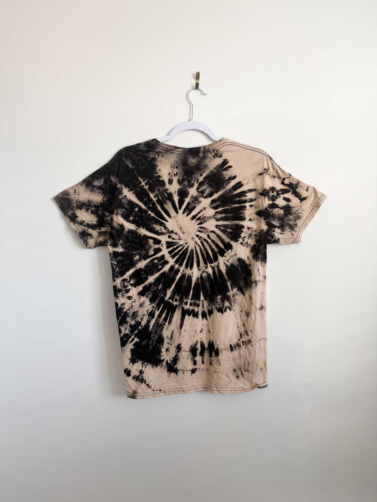 Back view of tan and black reverse spiral bleach tie dye t-shirt hanging on white hanger