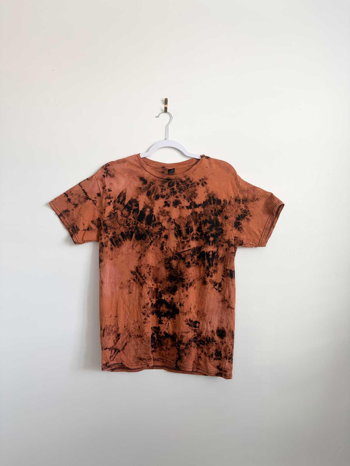 Medium rust orange and black bleach crumple tie dye t-shirt hanging on white hanger against white wall