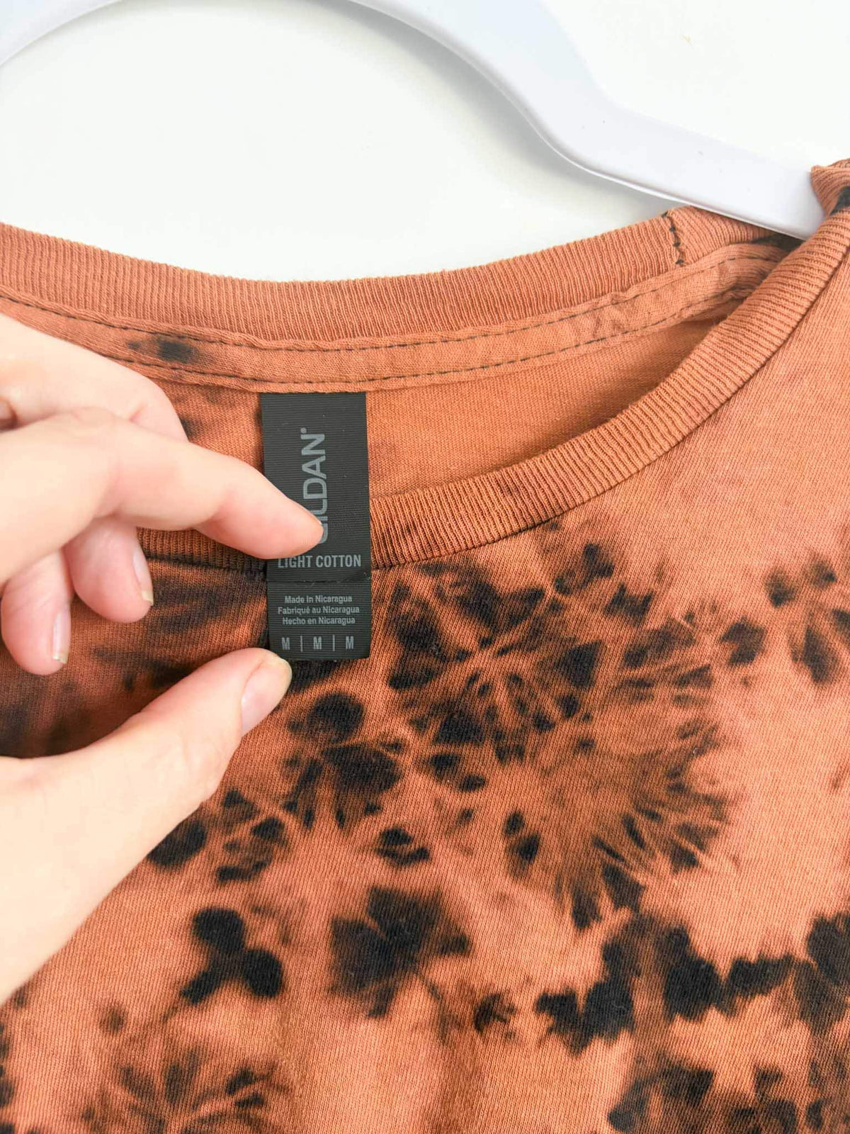 Close up of Gildan Light Cotton size medium tag on rust orange and black bleach crumple tie dye shirt