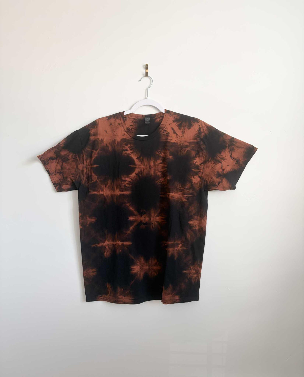 Large black and rust orange shibori bleach tie dye t-shirt hanging on white hanger against white wall