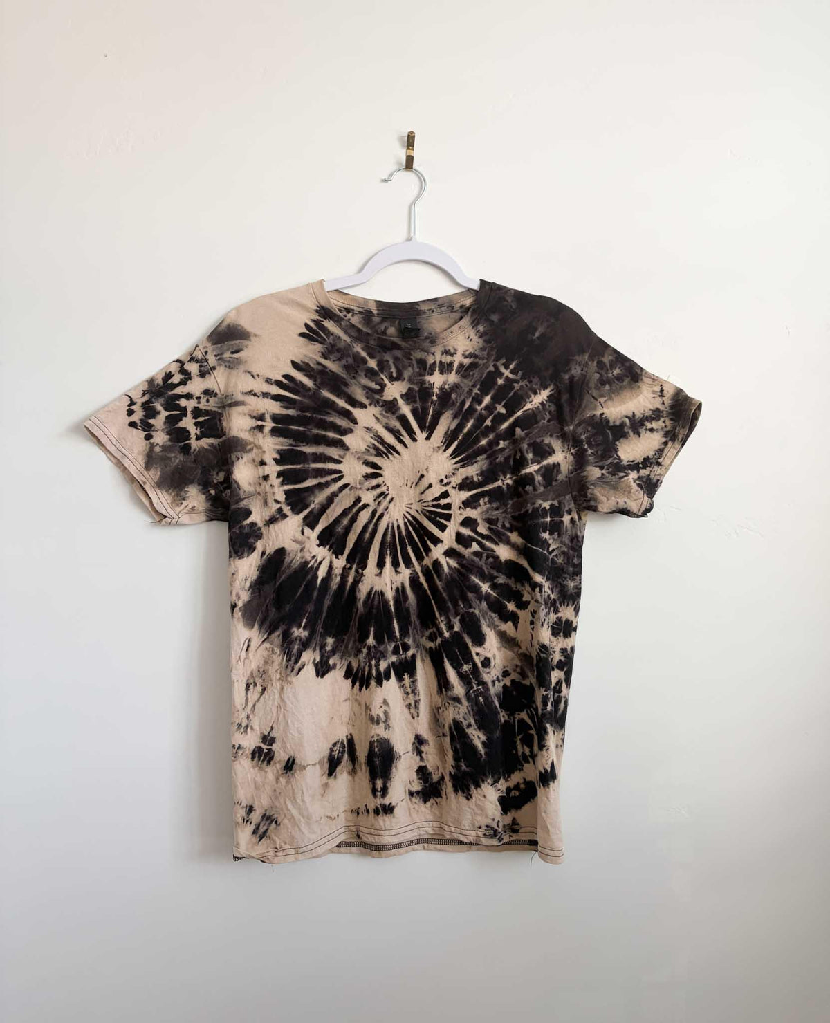 Medium tan and black reverse spiral bleach tie dye t-shirt hanging on white hanger against white wall