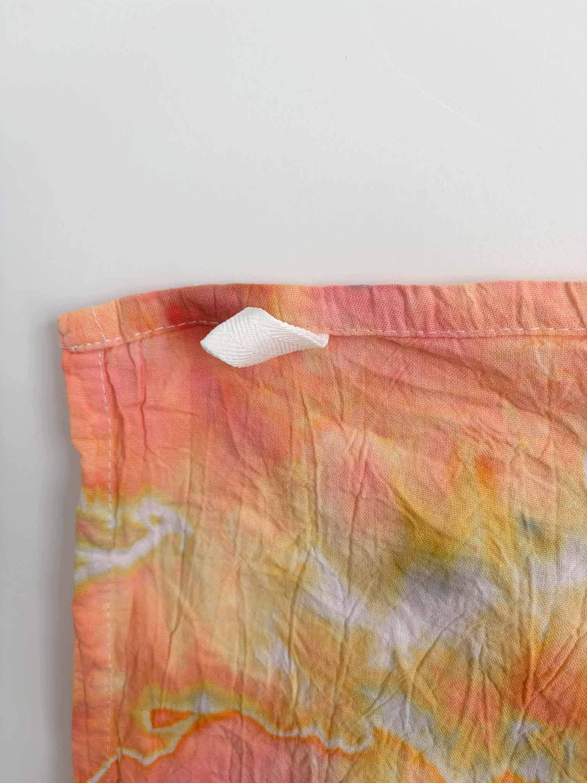 Corner of a coral and yellow ice dyed tea towel with a white twill hanging loop stitched into the hemmed edge.
