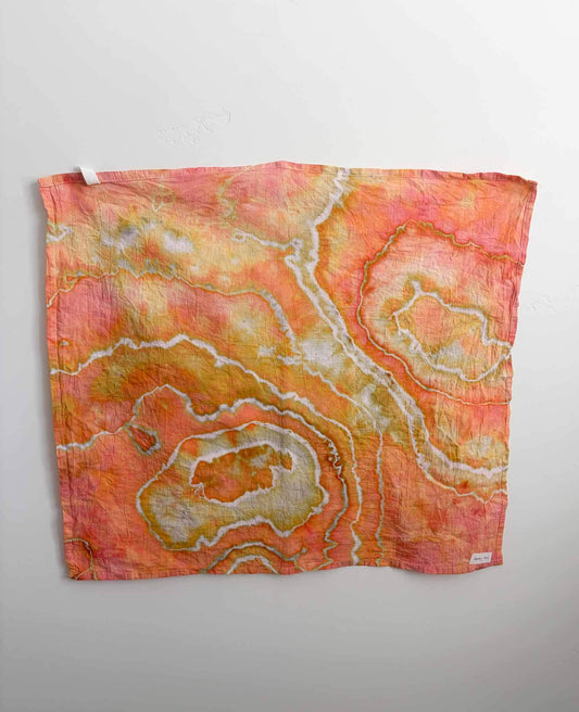 Square ice dyed tea towel hanging on a white wall, showing a geode pattern in coral, orange, and golden yellow with white crackle lines and olive green accents.