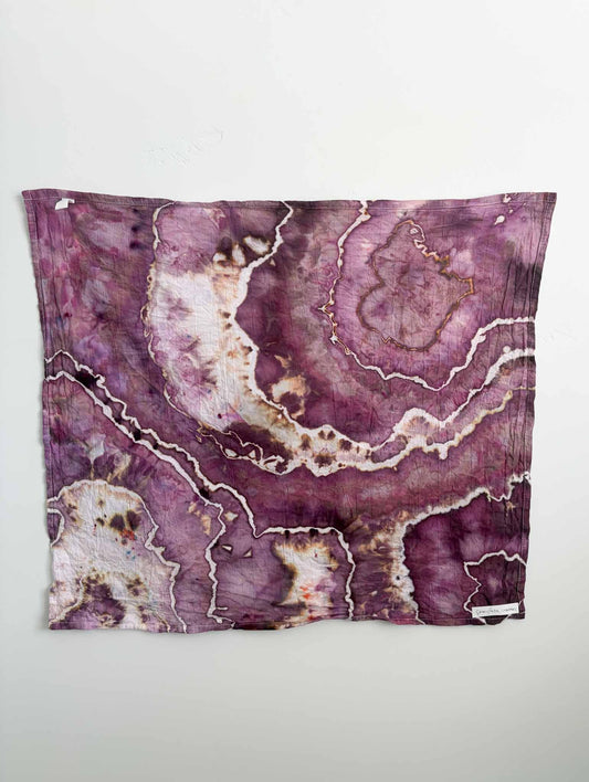 Square ice dyed tea towel hanging on a white wall, showing a geode pattern in berry plum purple with cream white bands, chocolate brown spots, and caramel accents.