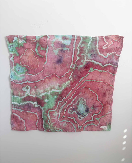 Square ice dyed tea towel hanging on a white wall, showing a geode pattern in raspberry pink with mint green and white crackle lines and purple accents.