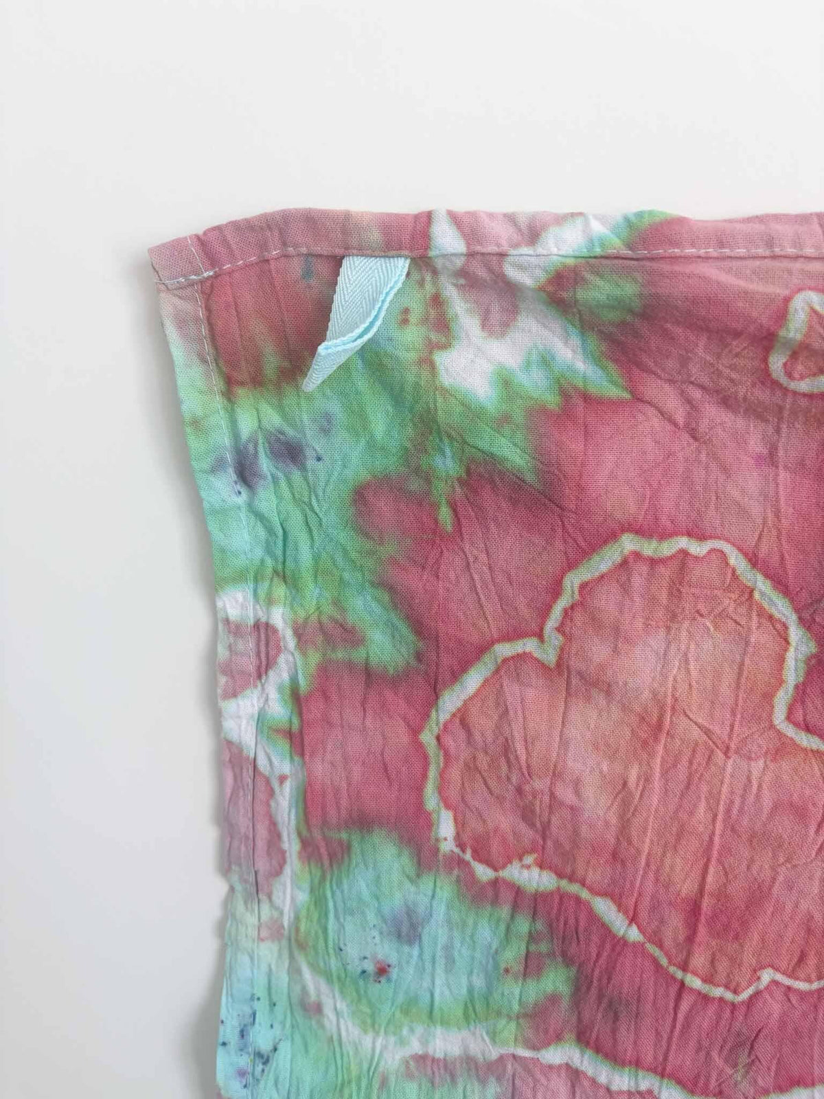 Corner of a pink and mint green ice dyed tea towel with a light blue twill hanging loop stitched into the hemmed edge.