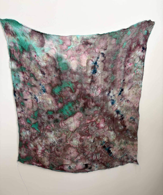 Square ice dyed bamboo gauze swaddle hanging on a white wall, showing a scrunch pattern in dusty rose, mauve, and berry with patches of jade green and navy blue.