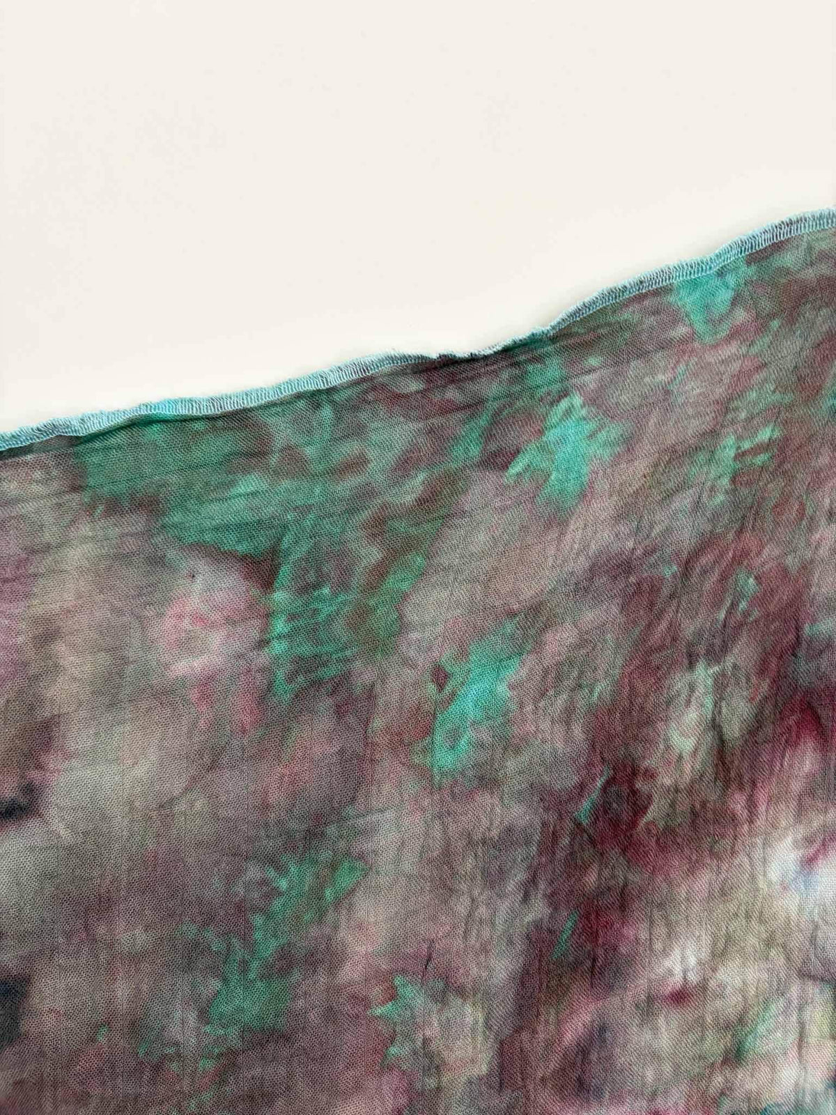 Edge of a rose and jade green ice dyed gauze swaddle showing the soft, sheer weave and an aqua overlocked hem.