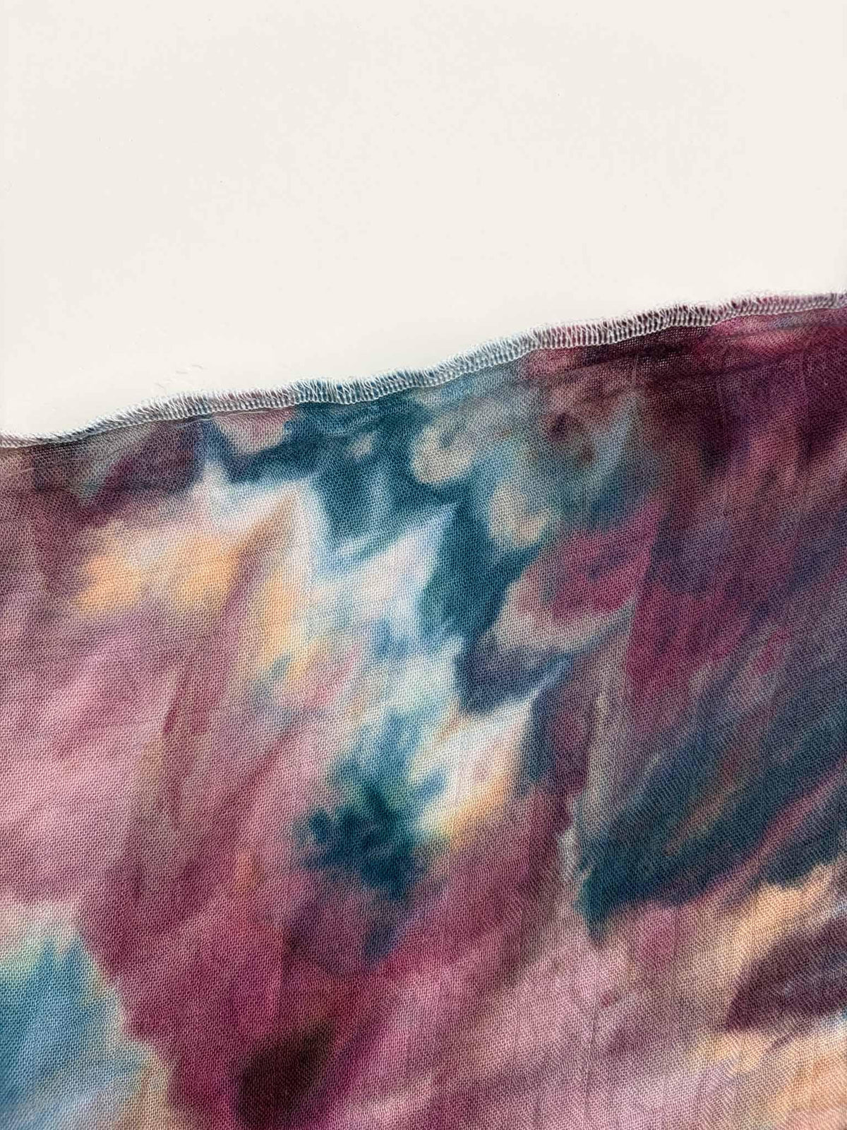 Edge of a berry and teal ice dyed gauze swaddle showing the sheer weave, soft peach patches, and a white overlocked hem.