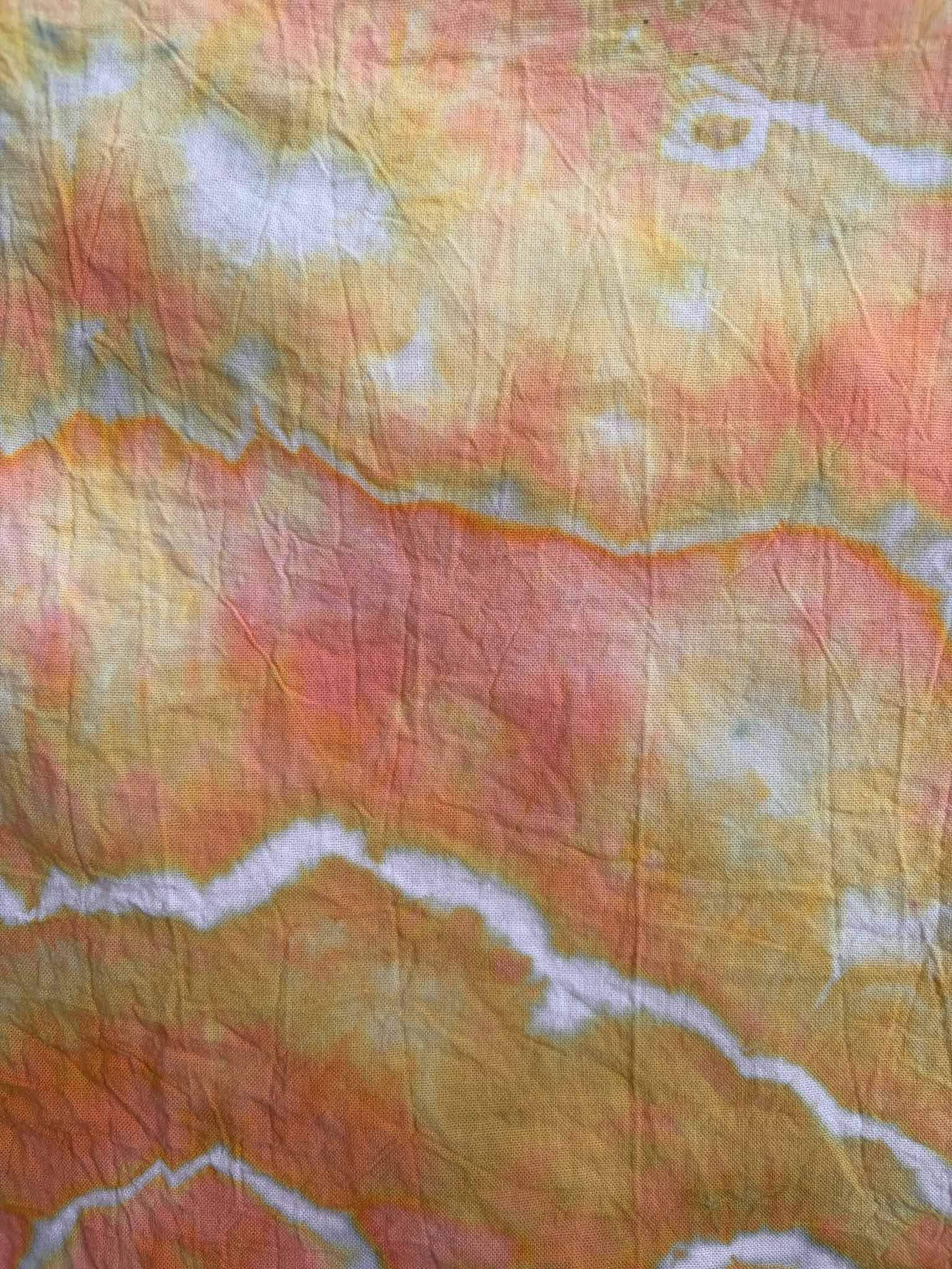 Close-up of ice dyed cotton showing coral and mustard yellow dye separating along jagged white lines, with soft green speckles where the colors blend.