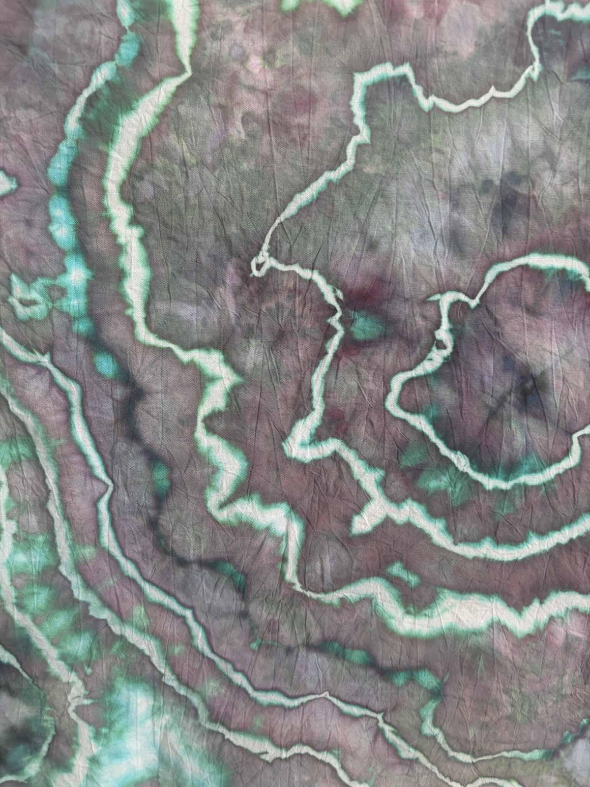 Close-up of ice dyed cotton with jagged white lines edged in teal green winding through soft plum and gray purple dye.
