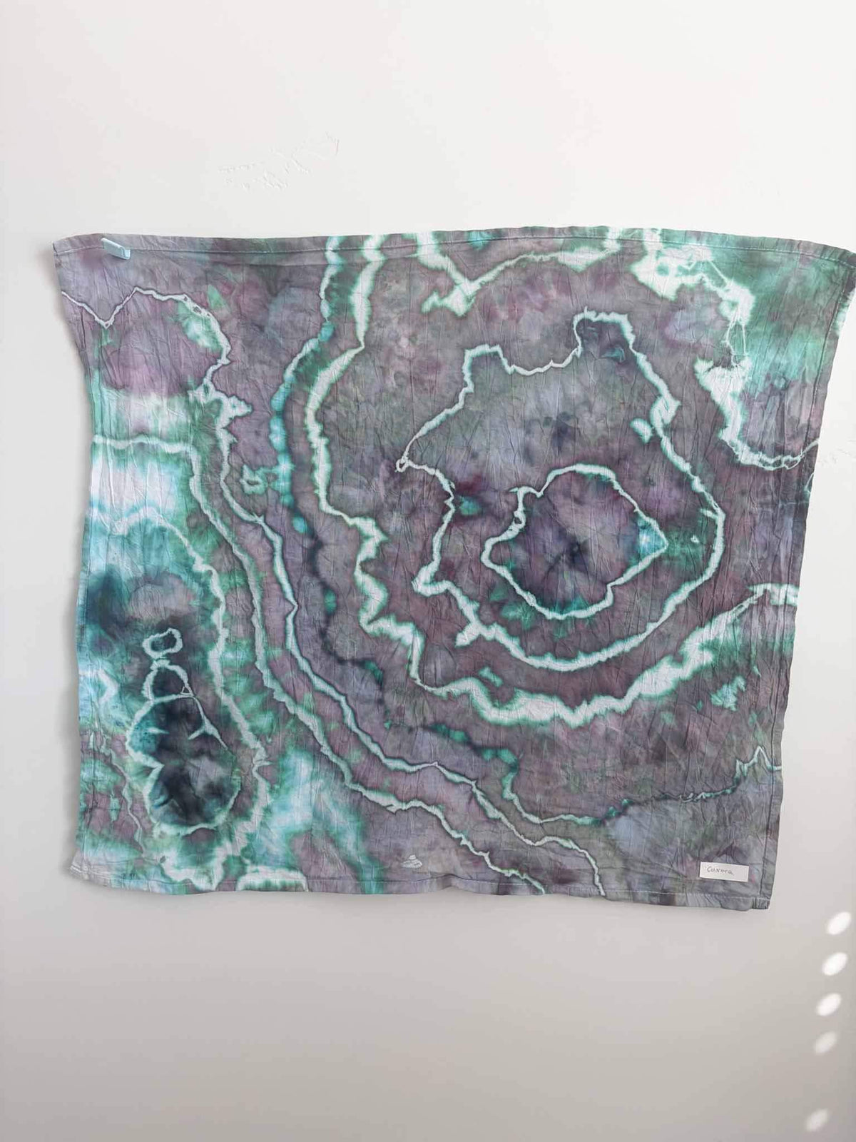 Square ice dyed tea towel hanging on a white wall, showing a geode pattern in dusty plum and mauve with teal green and white crackle lines and deep navy accents.