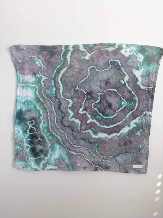 Square ice dyed tea towel hanging on a white wall, showing a geode pattern in dusty plum and mauve with teal green and white crackle lines and deep navy accents.