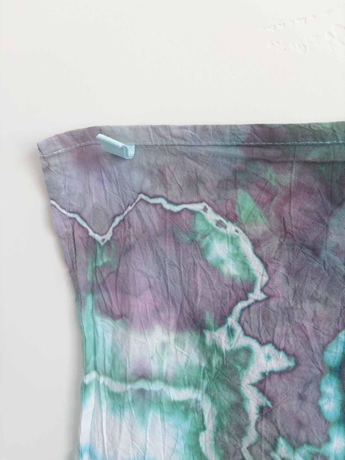 Corner of a purple and teal ice dyed tea towel with a light blue twill hanging loop stitched into the hemmed edge.