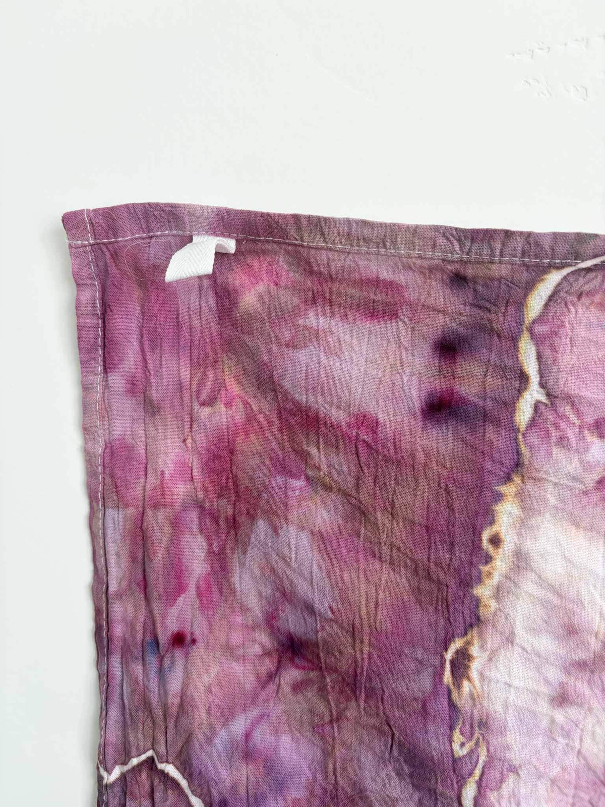 Corner of a plum and pink ice dyed tea towel with a white twill hanging loop stitched into the hemmed edge.
