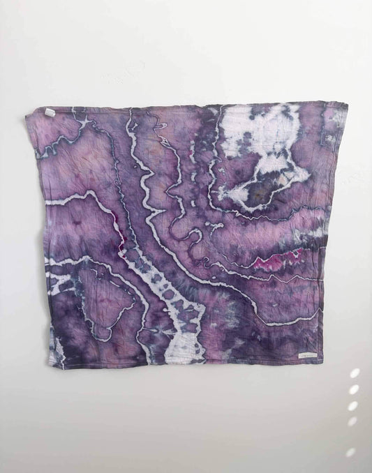 Square ice dyed tea towel hanging on a white wall, showing a geode pattern in violet and orchid purple with jagged white lines and bright magenta accents.